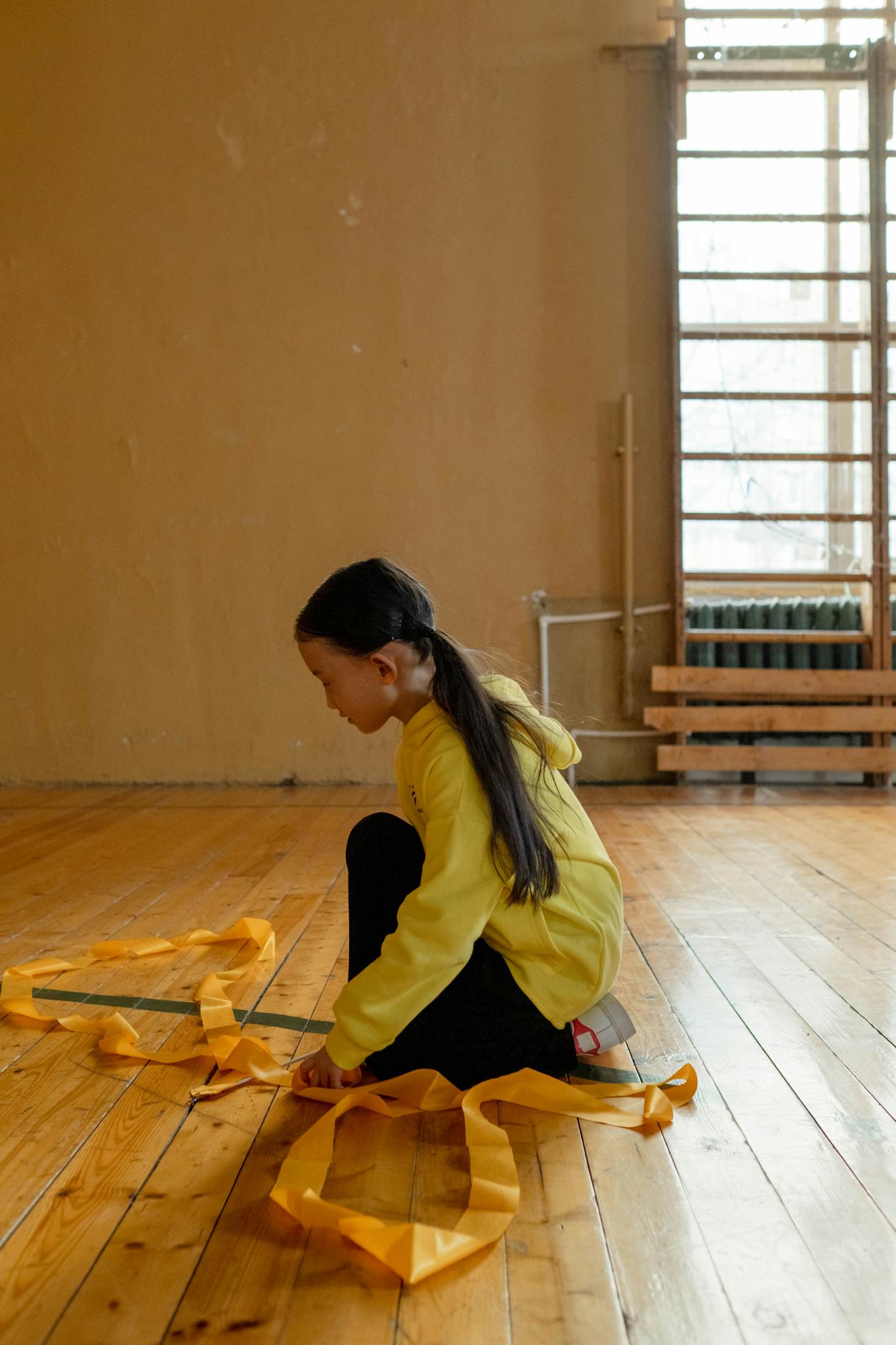 Child practicing gymnastics on a wooden floor indoors, wearing a yellow jacket, engaging in sports activity.